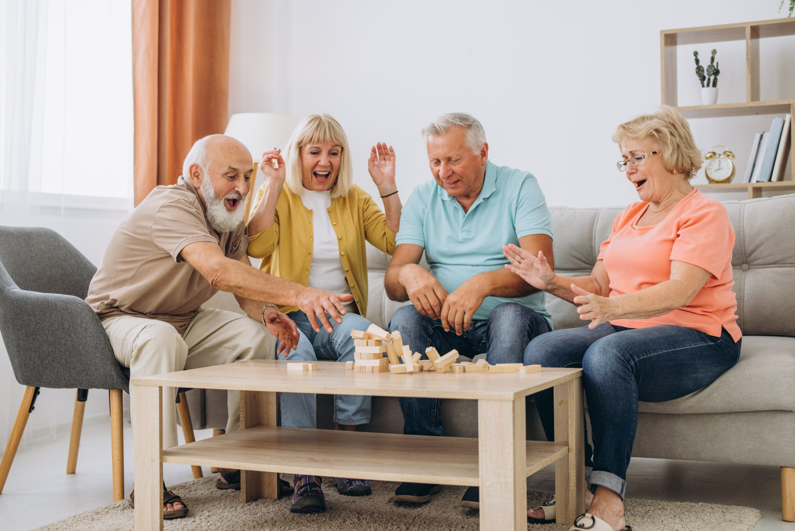 Group of happy senior people having fun and playing board games in retirement home. Several cheerful old men and women gather around table and take turns to take wood blocks out of wooden tower