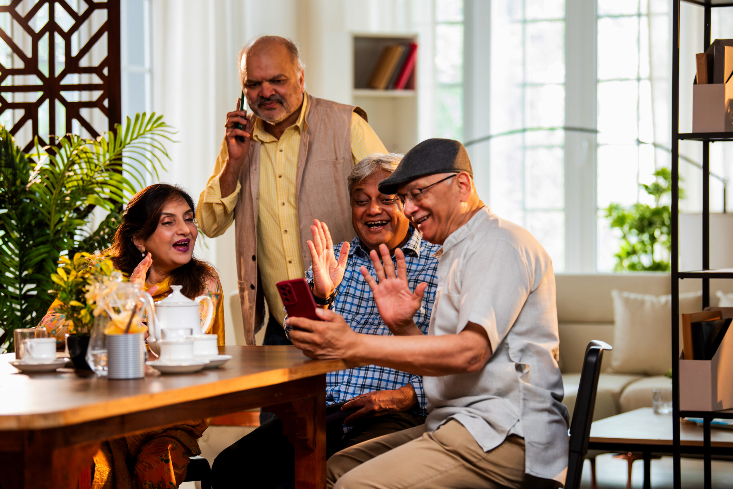 Seniors video calling at dining table, Indian Asian old age friends reuniting at home, attending group call using smartphone while enjoying tea, snacks, friendly talk at modern indoor dining setup
